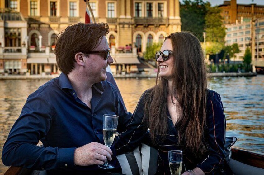 Two people sitting on a boat, holding glasses of a clear liquid, with a river and buildings in the background. 3