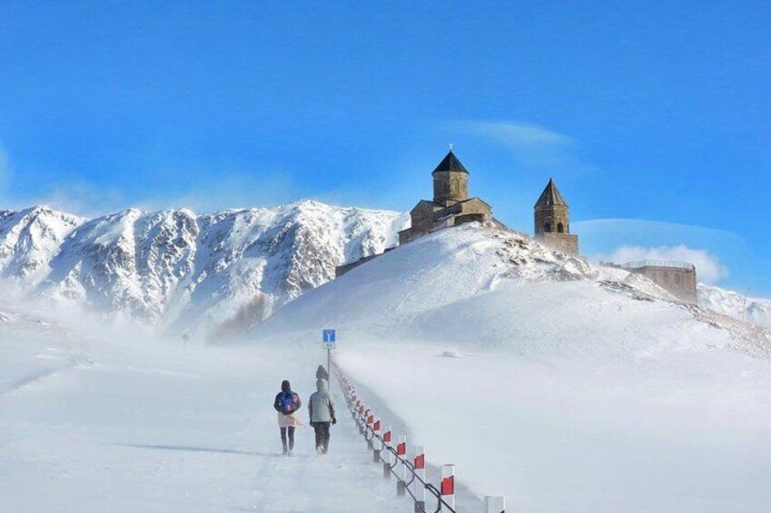 Camino nevado que conduce a un edificio histórico con dos torres, rodeado de montañas. 2