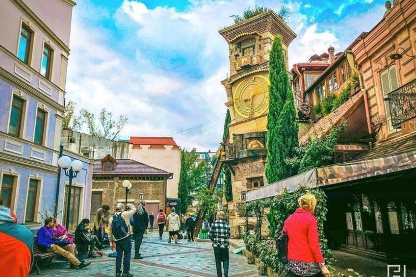 Una calle con gente caminando, una torre del reloj y edificios con balcones. 4