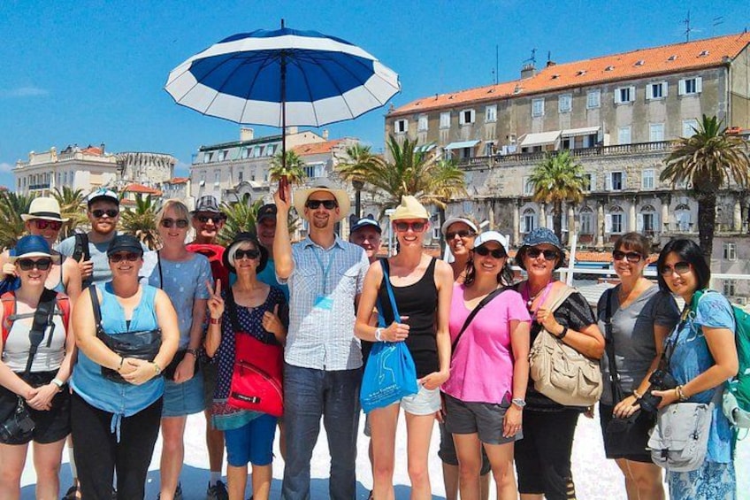 Een groep mensen staat op een zonnig strand, met een historisch gebouw op de achtergrond. 4