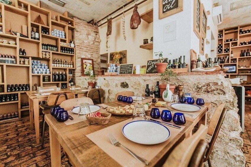 L'interno accogliente di una trattoria rustica, con scaffali di legno pieni di bottiglie, un lungo tavolo in legno apparecchiato per la cena e una parete di pietra sullo sfondo. 4