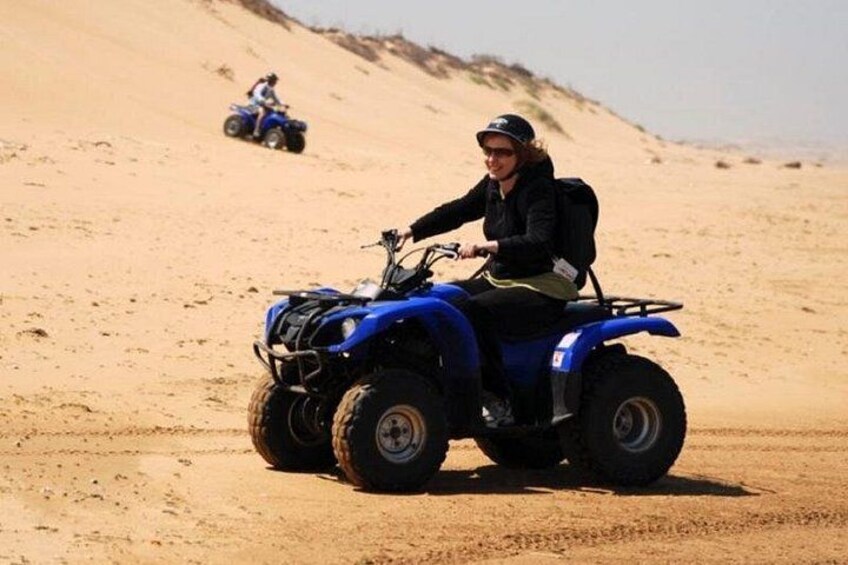 Iemand rijdt op een blauwe quad over zand met een duin op de achtergrond. 4