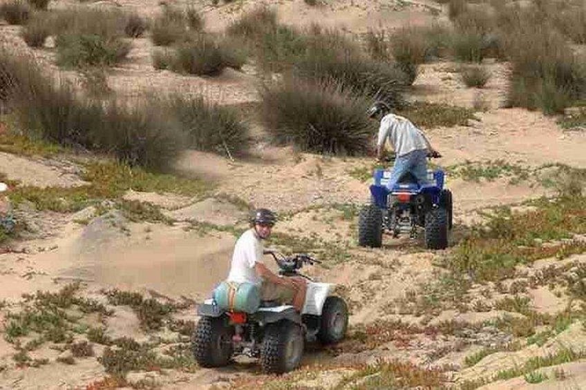 Twee personen rijden op quadbikes over een zandig terrein met weinig begroeiing. 2