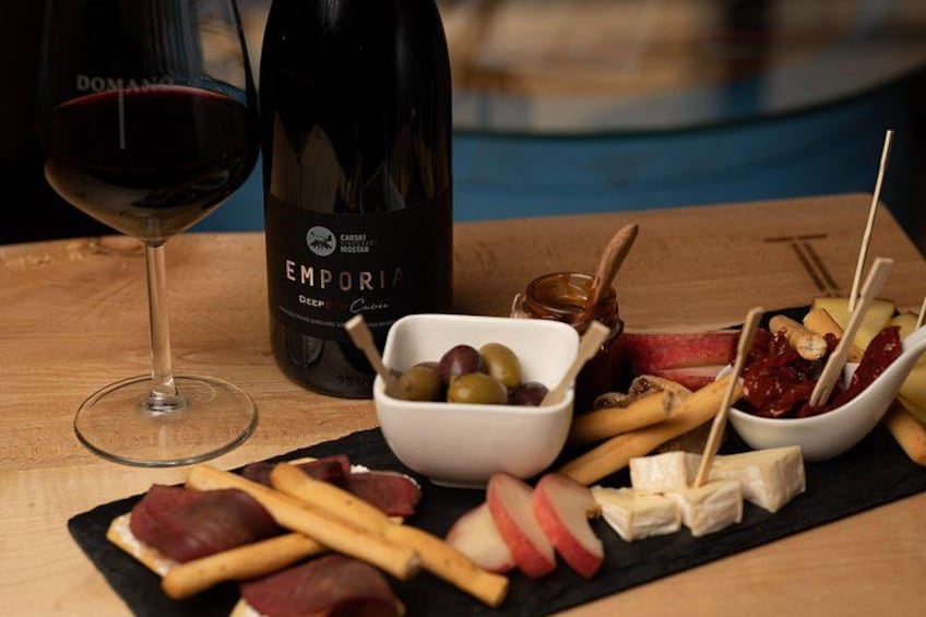 En flaske Emporia vin, et glas rødvin samt et udvalg af oste og frugter på et træbord. 2