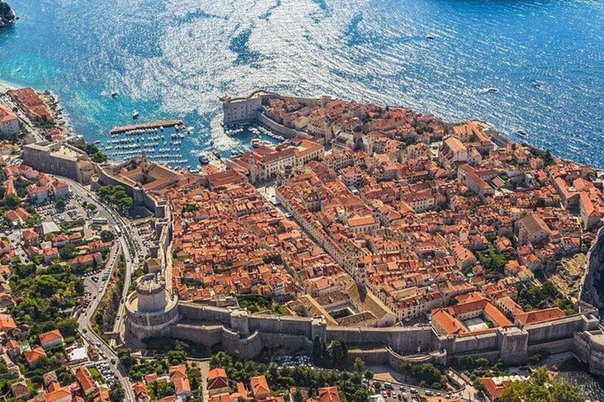 An aerial view of a coastal city with a prominent fortification, a dense urban area with red-roofed buildings, and a clear blue sea. 1