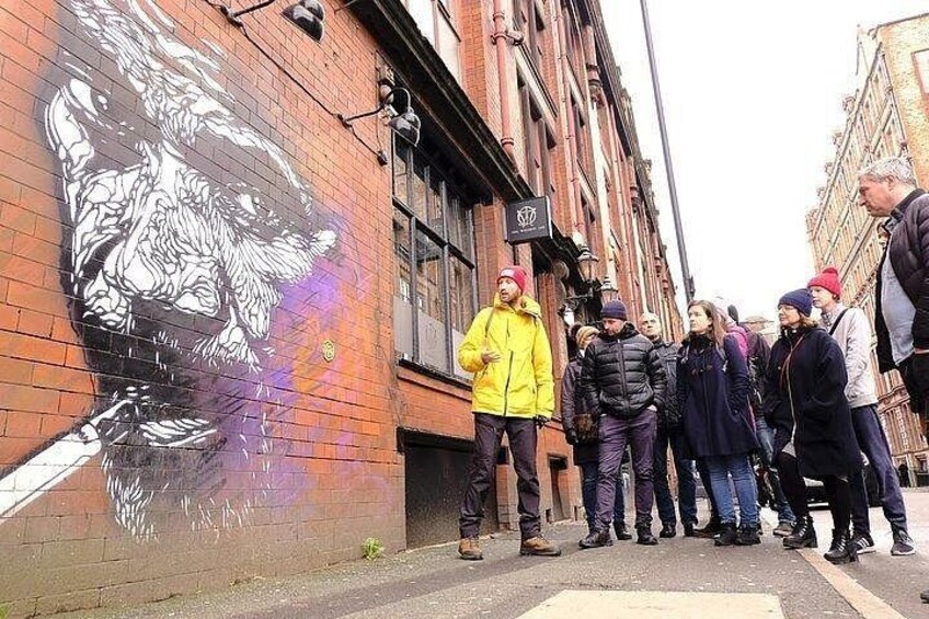 A mural on a brick wall, a group of people standing on the sidewalk, and a street with buildings. 2