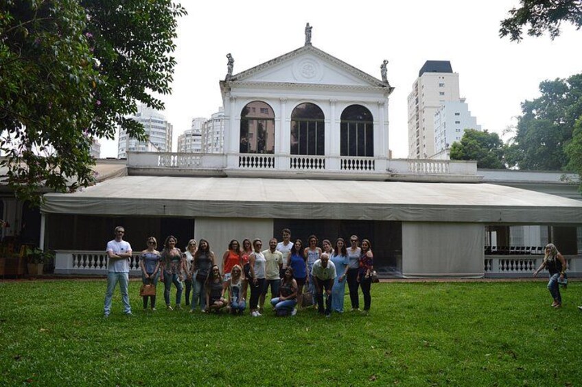 A group of people standing in front of a white building with a balcony and arched windows. 2