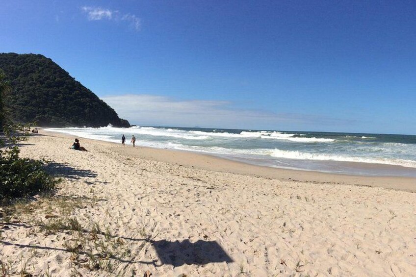 A sandy beach with waves, a person sitting, and a hill in the background. 2
