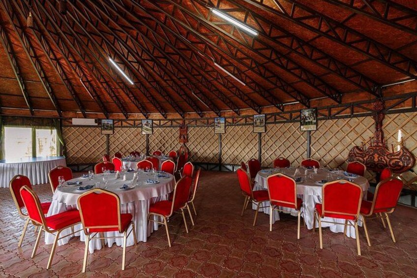 A large hall with red chairs and round tables, wooden beams on the ceiling, and a patterned wall. 5