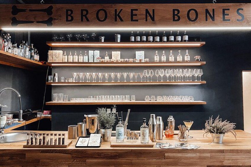 Eine Bar mit Regalen voller Flaschen und Gläsern, einer Holztheke und einem Schild mit der Aufschrift 'BROKEN BONES'. 5