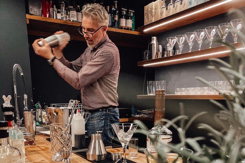 Eine Person mischt ein Getränk in einer Bar, im Hintergrund sind Regale mit Gläsern und Flaschen zu sehen. 1