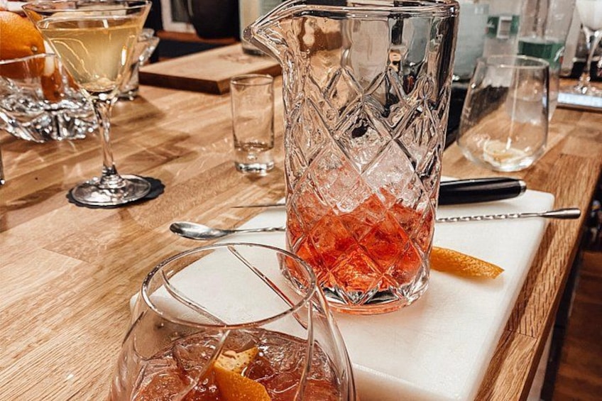 A bar setting with a pitcher of a red beverage, a glass with ice, and a glass of white drink on a wooden table. 2
