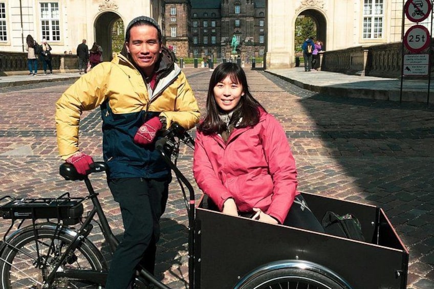 En person står bredvid en cykel med en kärra kopplad till den, på en utomhusplats i en stadsmiljö med stenlagd mark och historiska byggnader i bakgrunden. 3