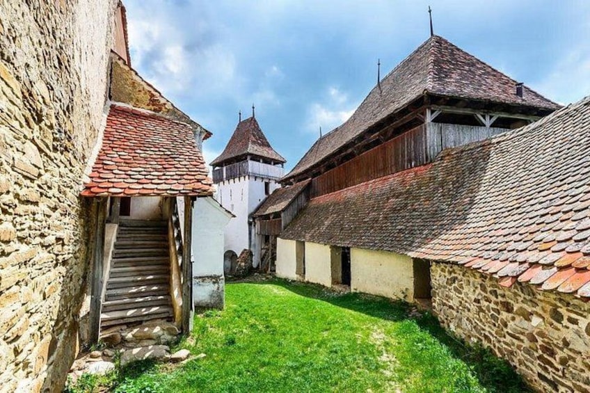 A medieval stone building with a wooden staircase, a grassy courtyard, and a tower with a conical roof. 2