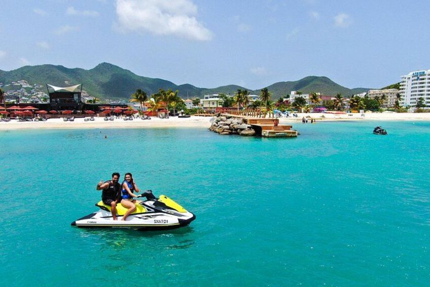 En jetski på turkisblåt vand med en strand og bjerge i baggrunden. 2