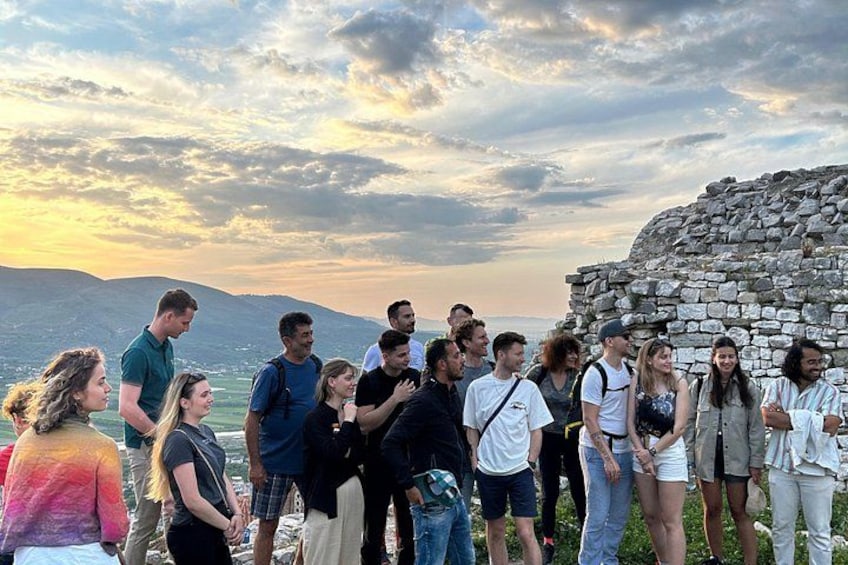 A group of people standing near a stone wall by a body of water with mountains in the background. 3