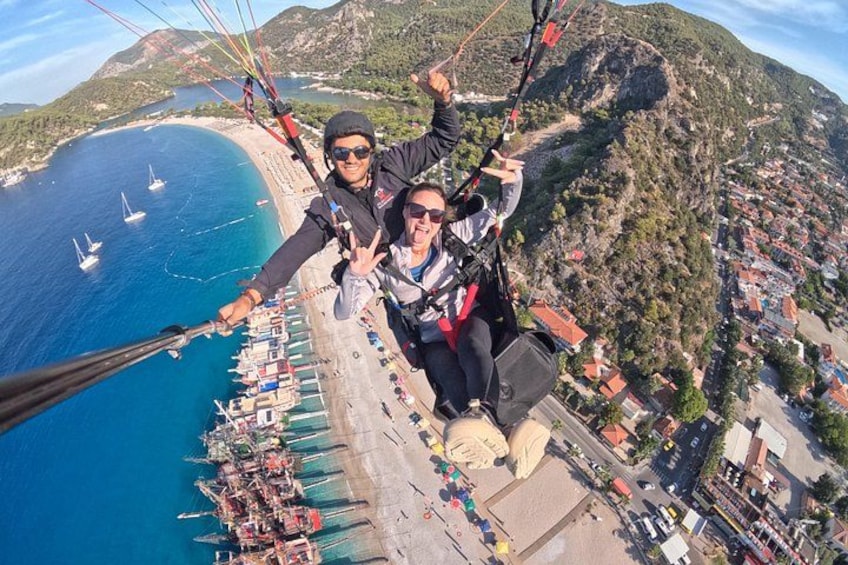 En person paraglider over et kystområde med strand, både og et bjergrigt landskab. 5