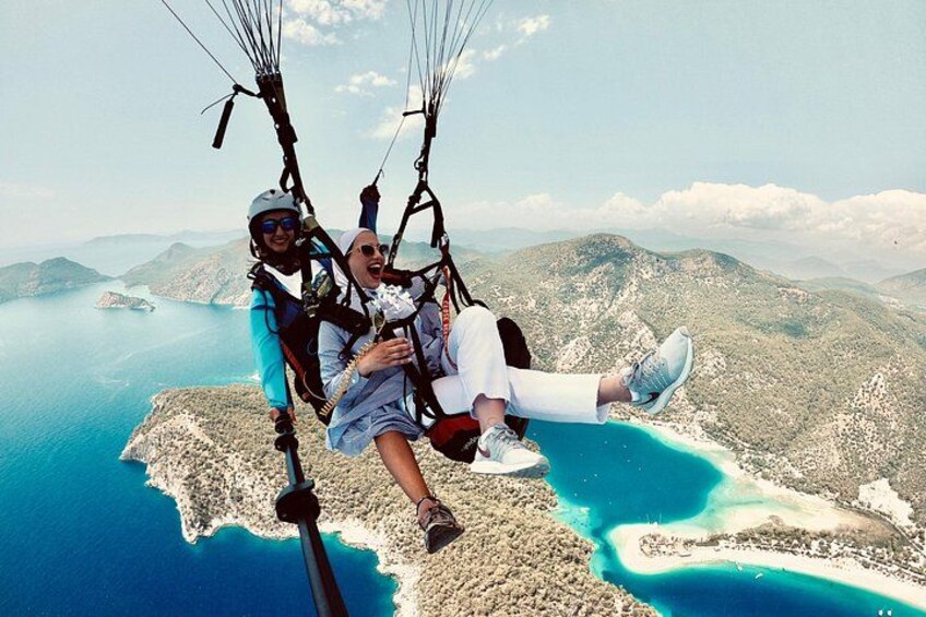 To personer paraglider over et kystlandskab med krystalklart, blåt vand og fjerne bjerge. 1