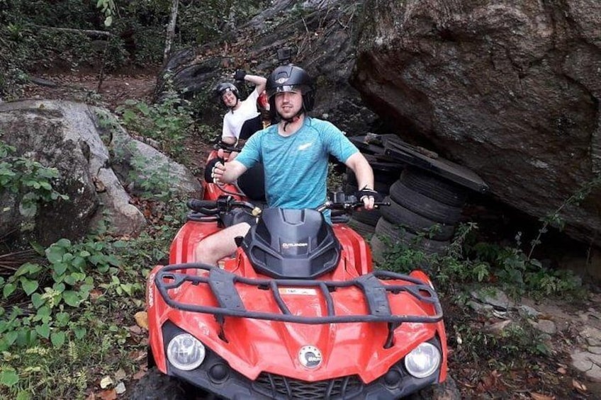 Un vehículo todo terreno rojo con dos personas en un área boscosa con rocas y follaje. 1
