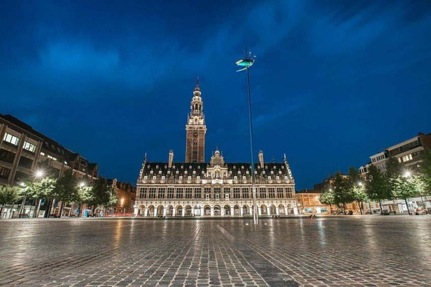 Un edificio histórico con una torre destacada, una plaza pavimentada y árboles circundantes. 2