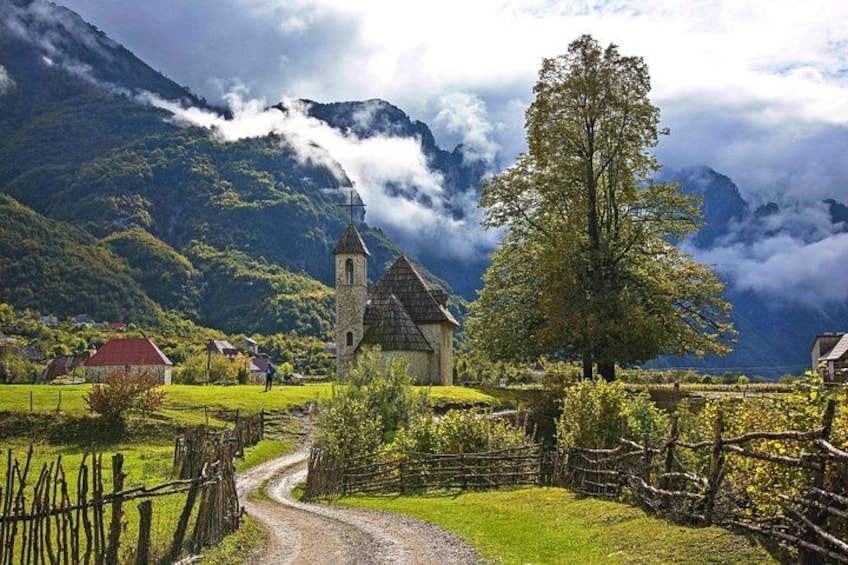 A rural landscape with a church, a large tree, and mountains in the background. 1