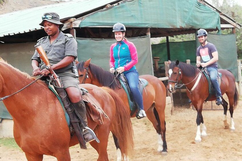 Three people riding horses in an outdoor setting with a dirt ground and a green-roofed structure in the background. 1