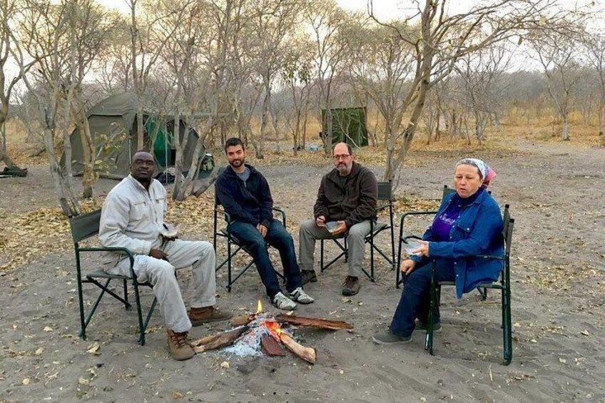 Four people sitting around a campfire in an open field with tents in the background. 2