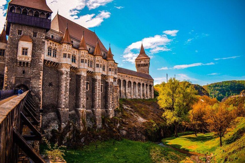 A historic castle with multiple towers, a bridge, and a lush autumn landscape. 1