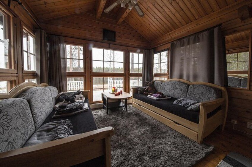 A cozy cabin interior with wooden walls, a sofa, a table, and a window overlooking a snowy landscape. 3
