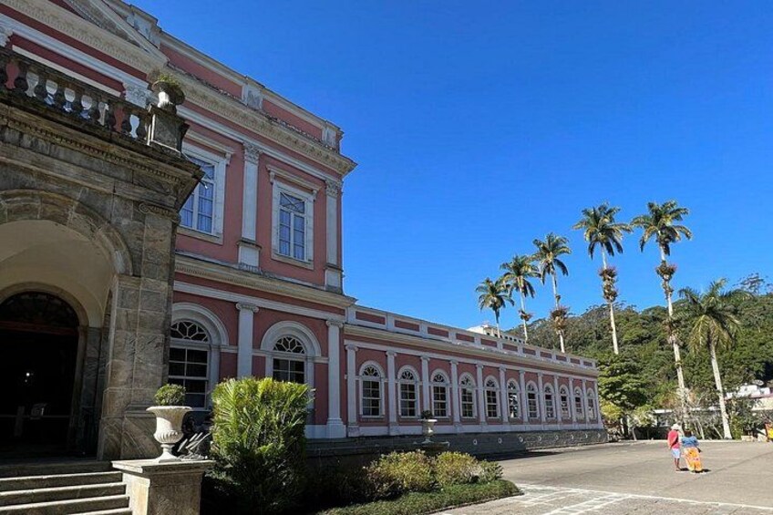 A historic building with arched windows and a pink facade, palm trees in the background, and a clear blue sky. 1