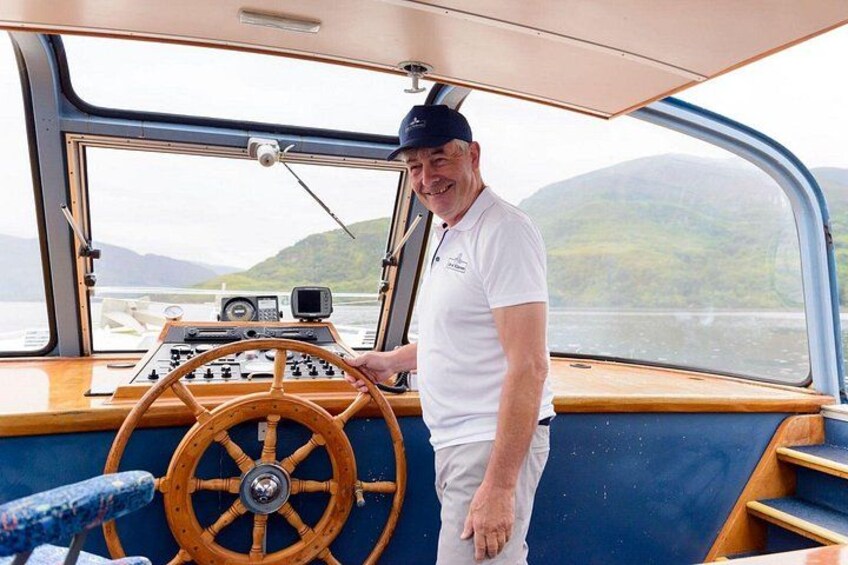 A man standing at the helm of a boat, steering wheel in hand, with a view of a calm lake and hills in the background. 2