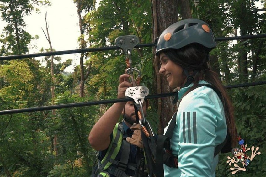 A person wearing a helmet and harness, holding a safety device, in a forested area with trees and a rope bridge. 2