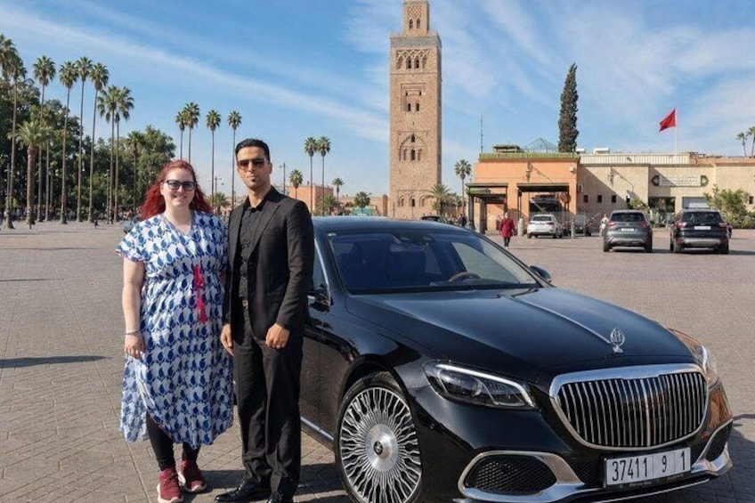 A black car, a woman in a blue dress, and a man in a suit standing in front of a building with a tower. 1