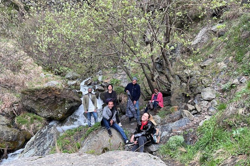 A group of people sitting and standing on rocks near a stream in a forested area. 2