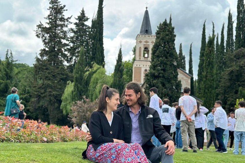 A couple sitting on grass with a church and tall trees in the background. 4