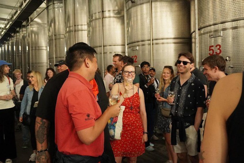 A group of people in a wine cellar with stainless steel tanks, some holding glasses. 3