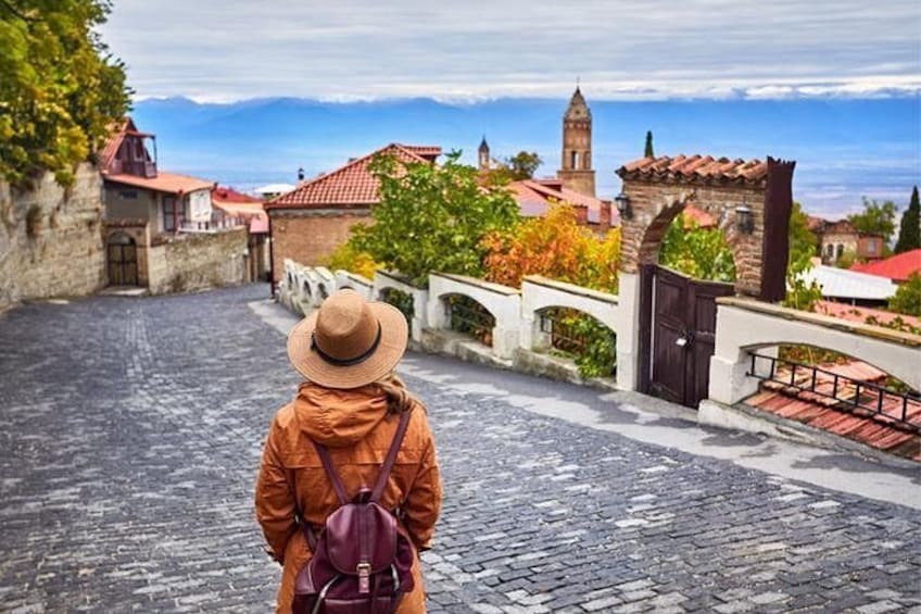 Una persona con chaqueta y sombrero marrón camina por una calle adoquinada, con vistas a un pueblo histórico con edificios y una torre. 1