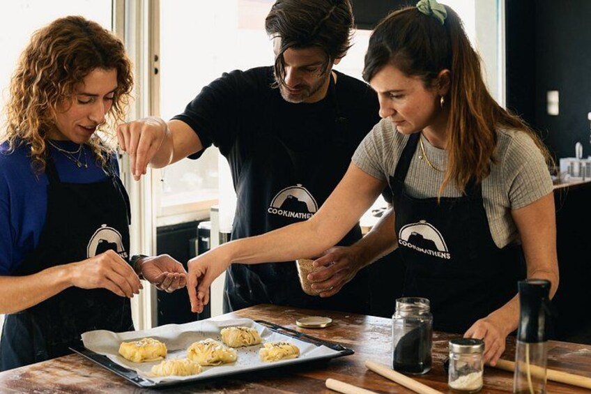 Drie mensen in schorten met een logo bereiden eten voor op een tafel met een bakplaat, deegroller en potten. 1