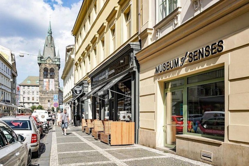 Een straat met geparkeerde auto's, een museum genaamd 'Museum of Senses' en een kerktoren op de achtergrond. 2