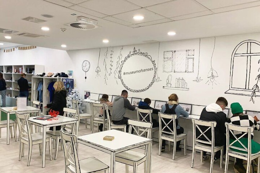 Ein Museumscafé mit Tischen und Personen, einer weißen Wand mit Skizzen und einem Hashtag-Symbol. 2