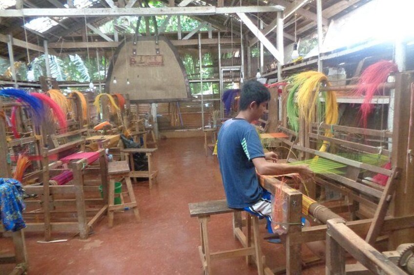 A person weaving colorful threads on a loom in a rustic workshop with wooden structures and a thatched roof. 1