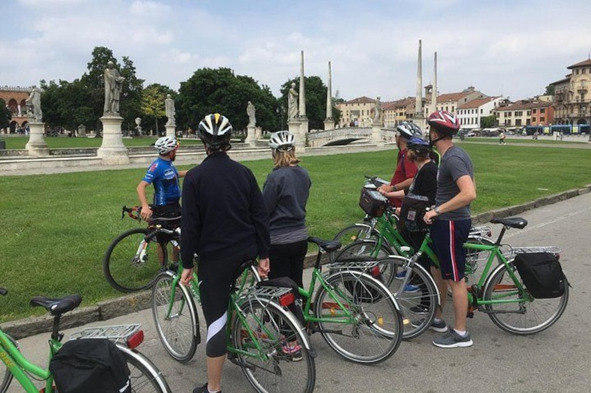 En gruppe cyklister samlet omkring cykler i en park med statuer og bygninger i baggrunden. 3