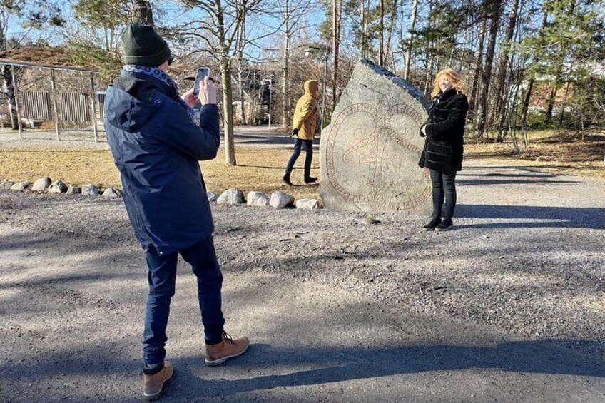 A person taking a photo of a large stone monument with inscriptions, surrounded by a gravel area and trees. 2