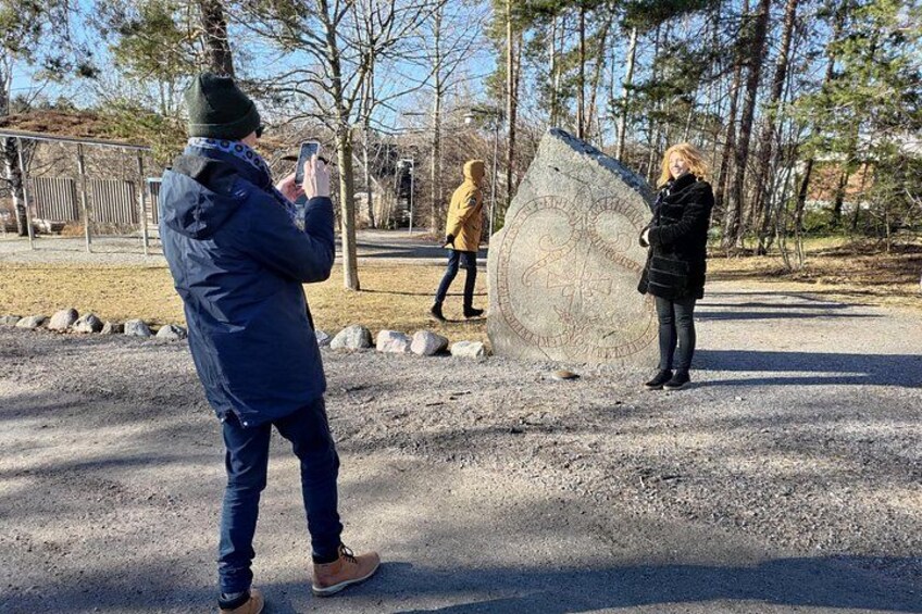 A person taking a photo of a large stone monument with inscriptions, surrounded by trees and a gravel path. 4
