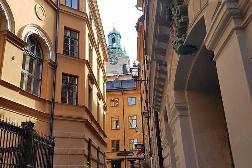 A narrow street with buildings, a clock, and a tower with a clock. 1