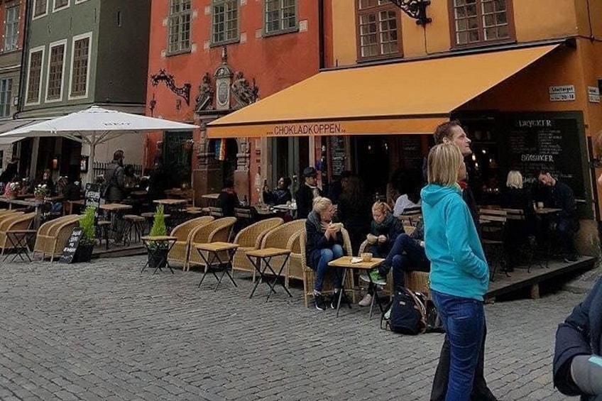 Outdoor seating with people dining, a building with a sign reading 'CHOKLADKOPPEN,' and a cobblestone street. 3