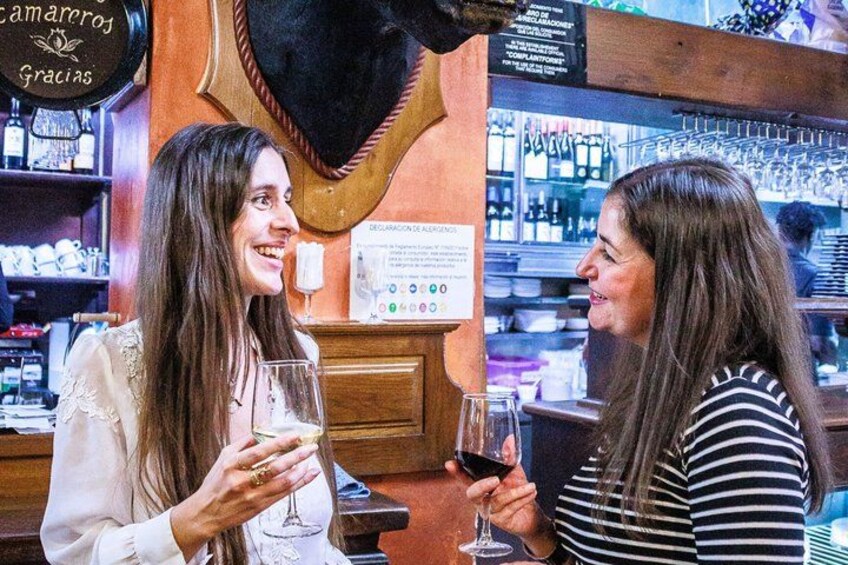 Two women at a bar with wine glasses, shelves with bottles, and a menu board. 3