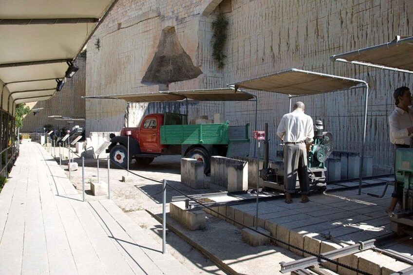 A construction site with a green truck, a man standing by a machine, and a large bell on a wall. 2