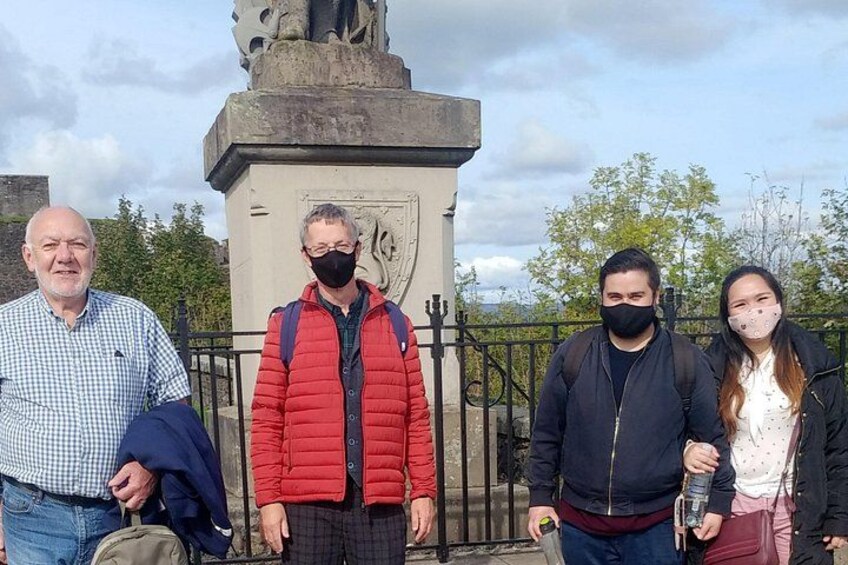 Vier personen staan voor een monument, met een hek en bomen op de achtergrond. 3