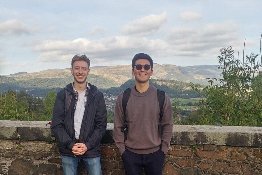 Two people standing on a stone wall with a mountainous landscape in the background. 3